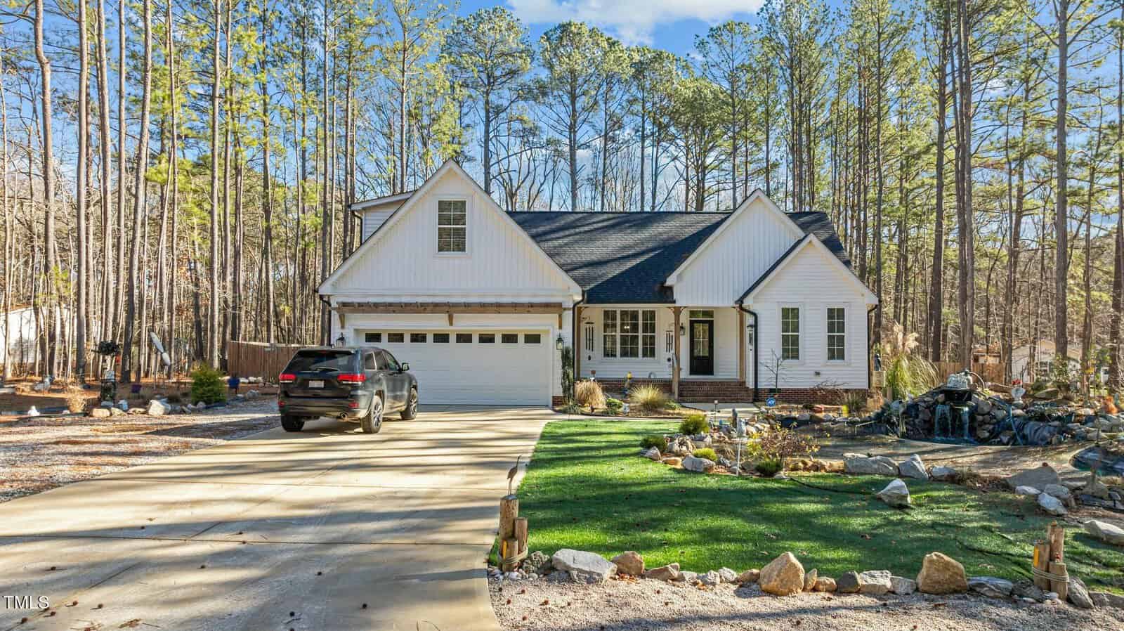 A white, single-story house with a black roof and attached garage sits among tall trees. A black SUV is parked in the driveway, and there is a landscaped yard with rocks and a small pond feature in front.