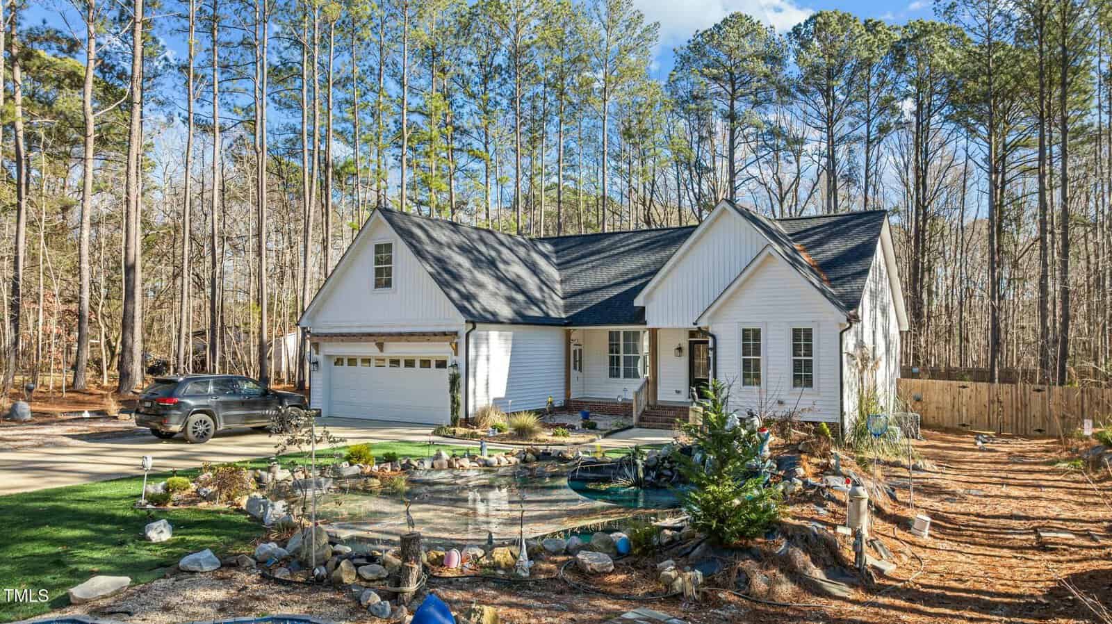 A white single-story house with a black roof sits in a wooded area. A black car is parked in the driveway, and a landscaped pond with rocks and plants is in the front yard. Tall trees surround the property.