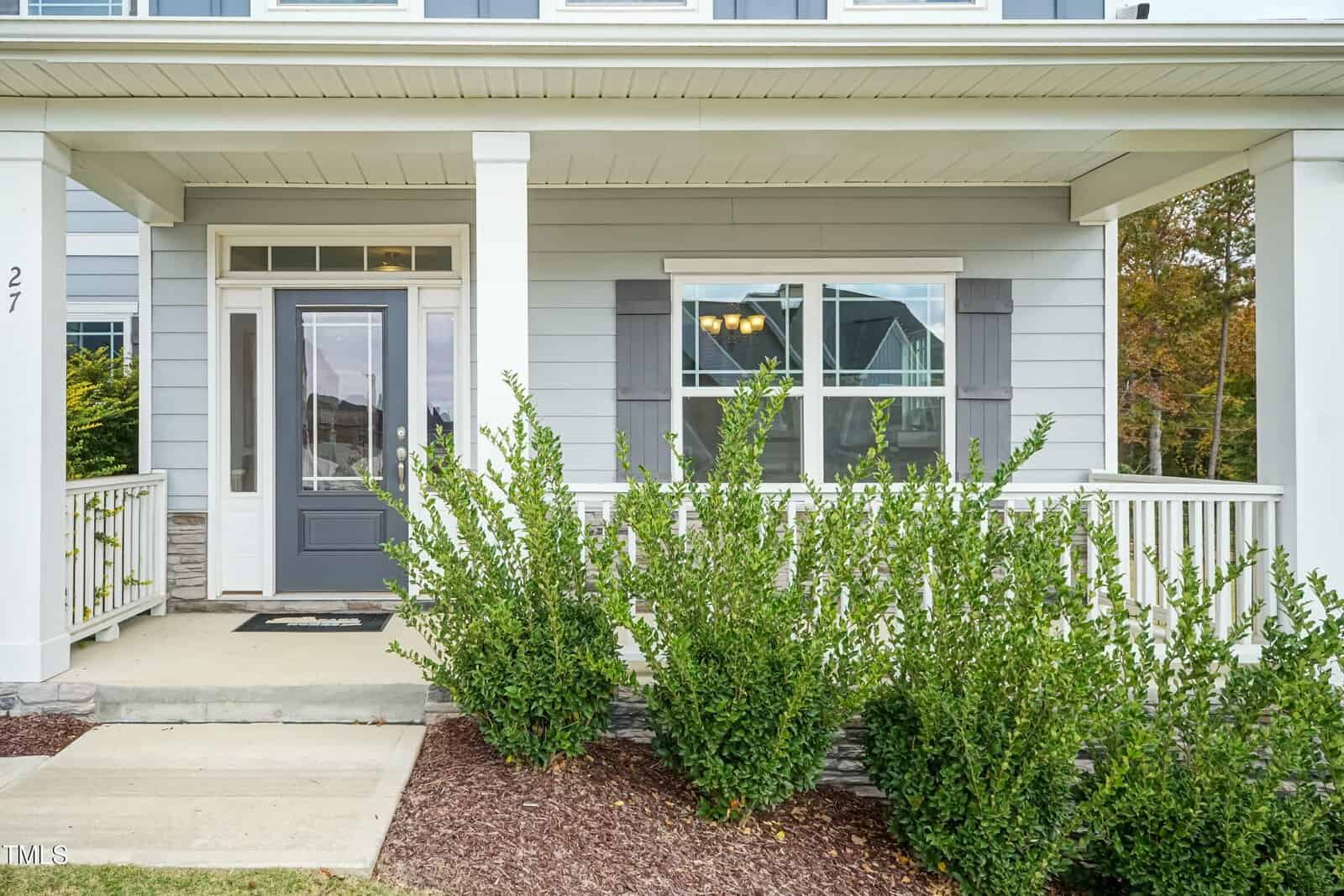 A gray front porch with white trim and railing, a dark gray front door with glass panels, two front-facing windows with shutters, and green shrubs planted along the walkway. House number 27 is visible on a column.