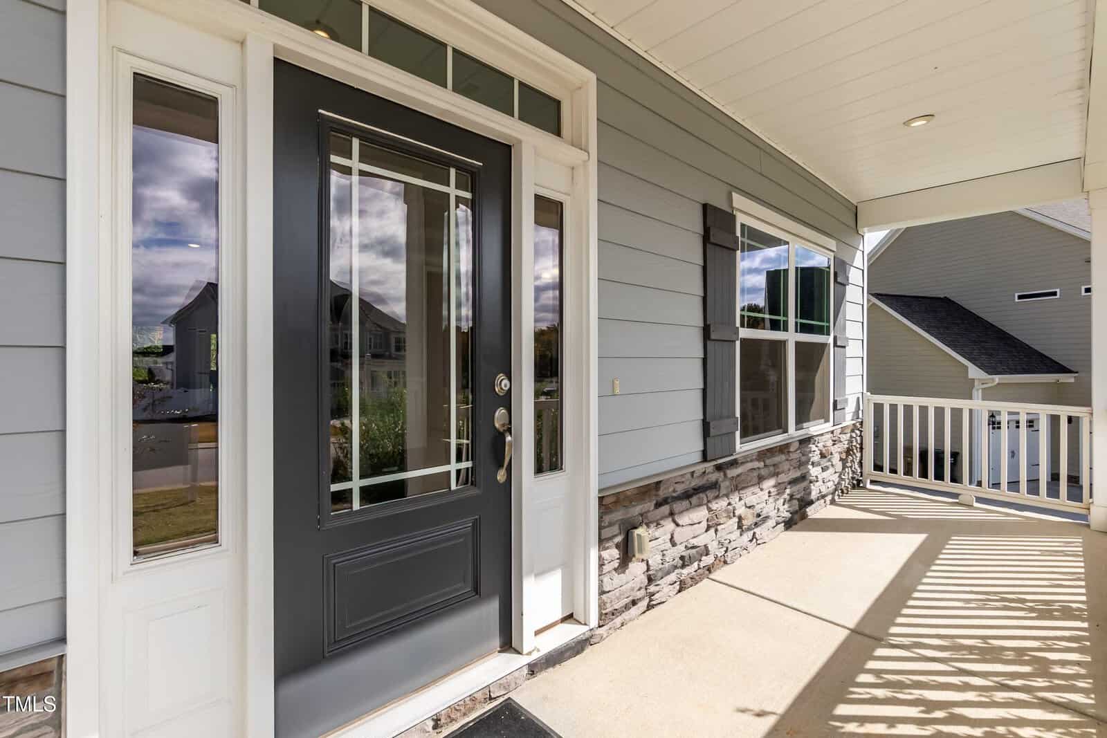 View of a front porch with a black front door featuring glass panels, sidelights, and a gray house exterior with stone accents and a white railing. Sunlight casts shadows across the porch floor.