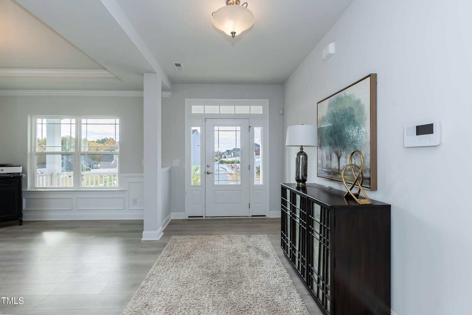 Bright entryway with double front doors, a rug on light wood flooring, a dark console table with a lamp and décor, and a large framed picture on a pale wall. Windows and white trim are visible to the left.