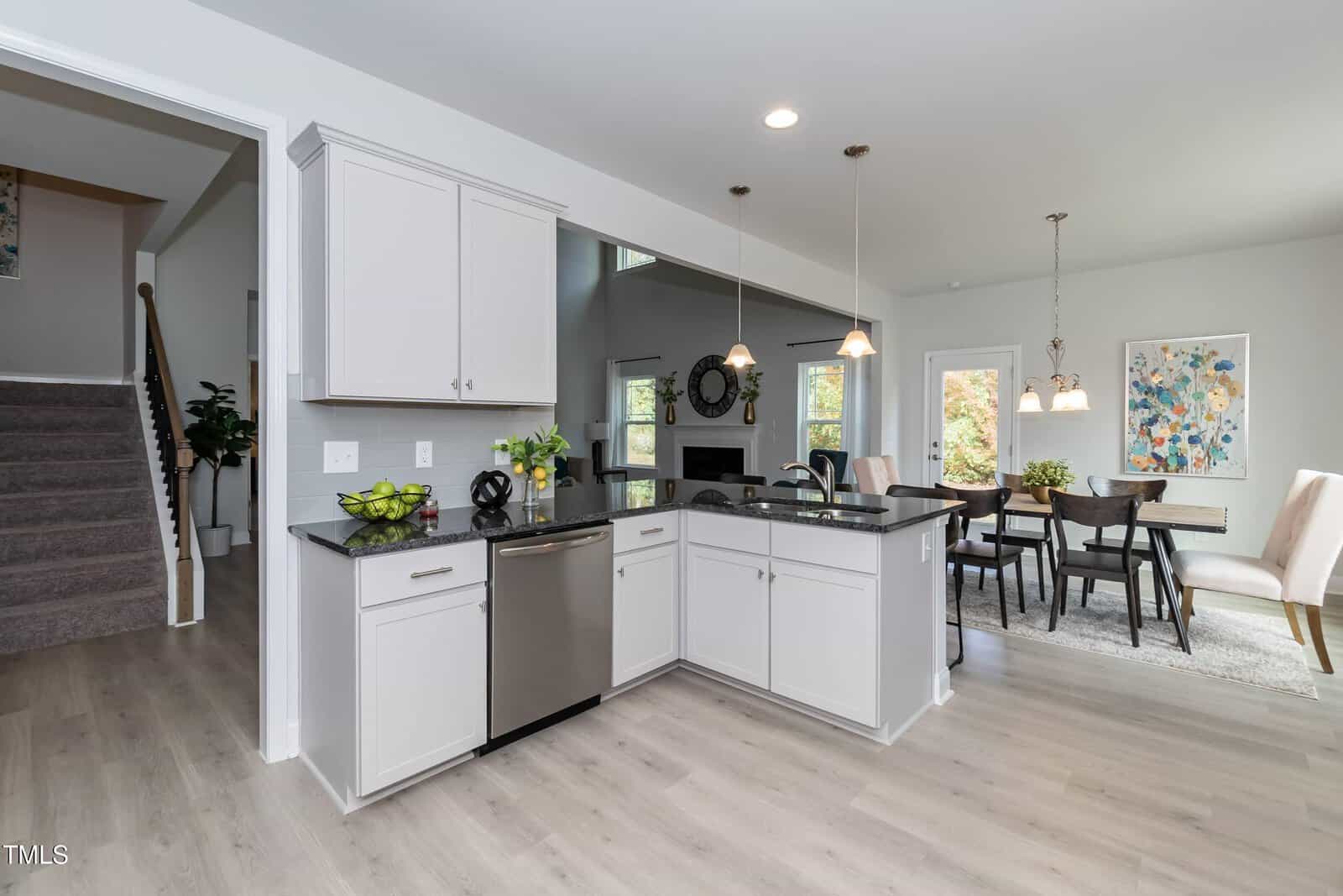 Modern kitchen with light cabinets, stainless steel appliances, and black countertops. Open layout connects to dining and living areas. Pendant lights hang above the counter, and a carpeted staircase is visible in the background.