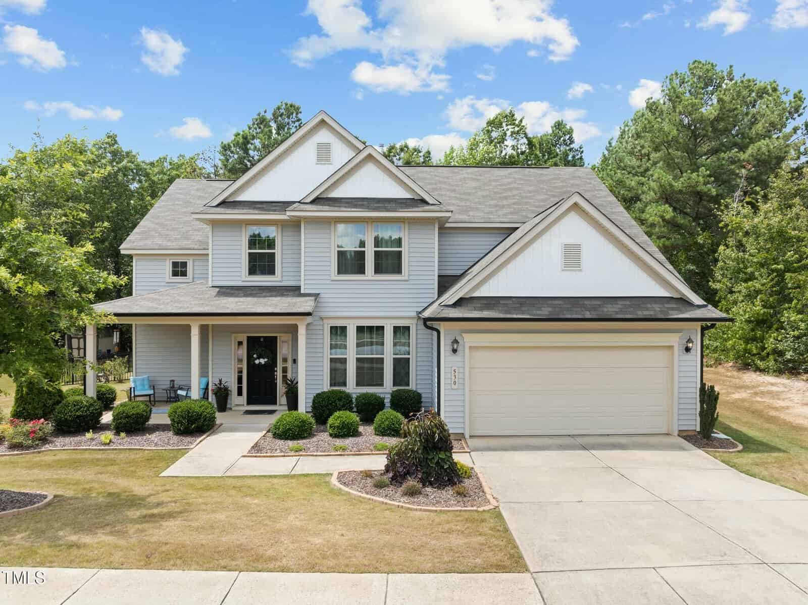 Two-story suburban house with light gray siding, white trim, and a two-car garage. The front yard has neatly trimmed bushes, small trees, and patches of grass bordered by mulch beds. A porch with seating is visible.