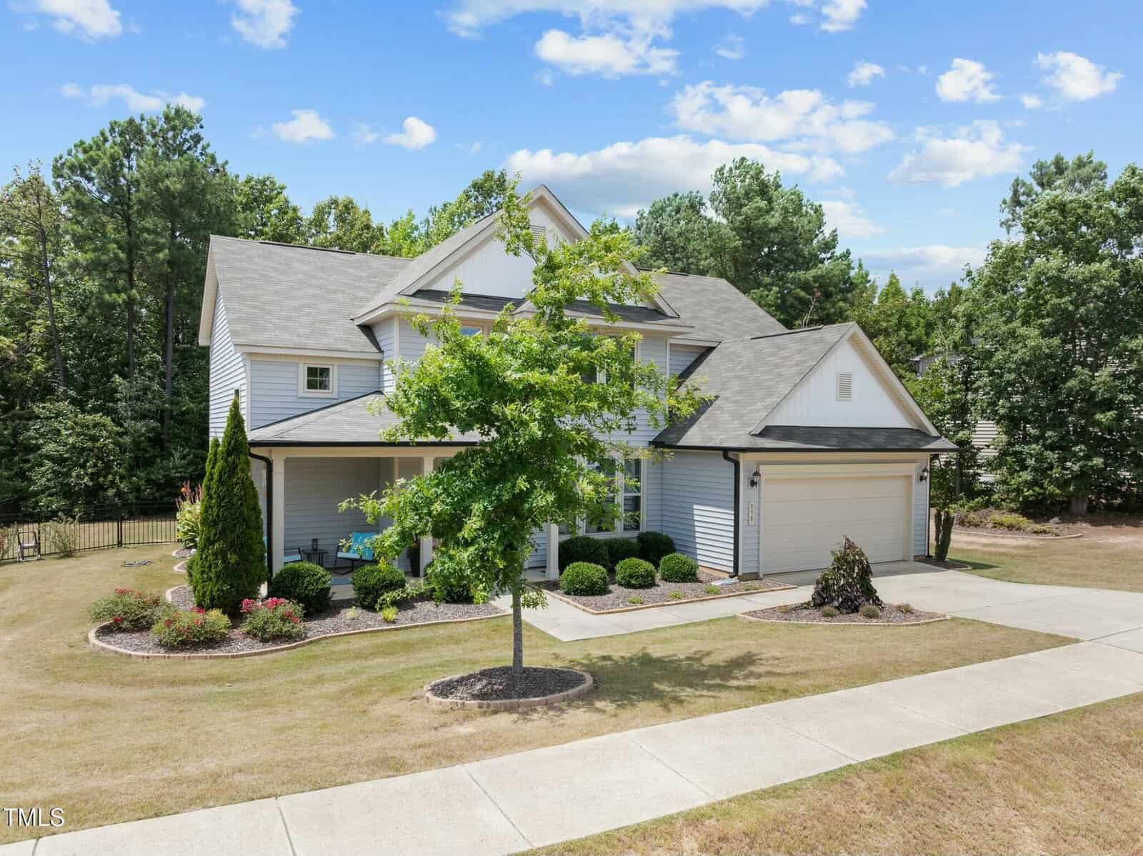 A two-story suburban house with light gray siding, white trim, and a covered front porch sits on a neatly manicured lawn with shrubs and a small tree. The driveway leads to an attached two-car garage.