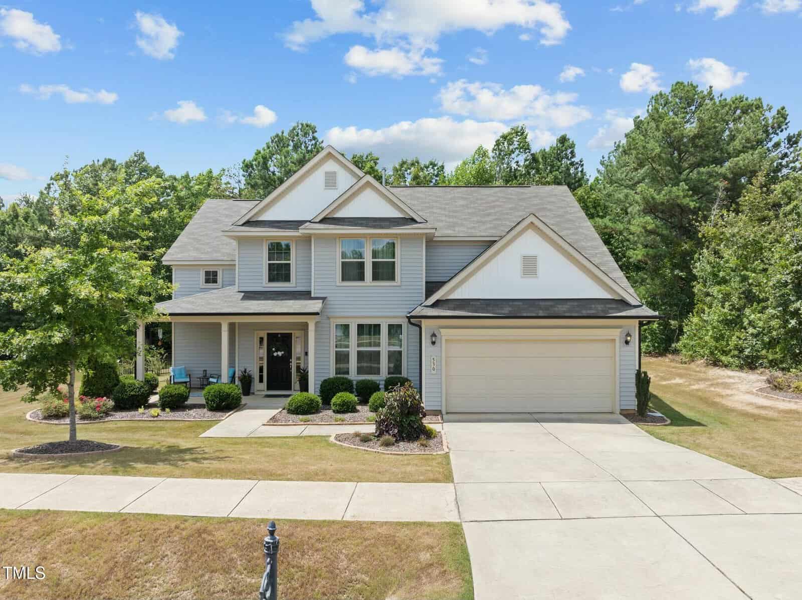 A two-story suburban house with gray siding, white trim, a covered front porch, and an attached two-car garage. The home is surrounded by bushes and trees, with a driveway and lawn in front, and woods in the background.