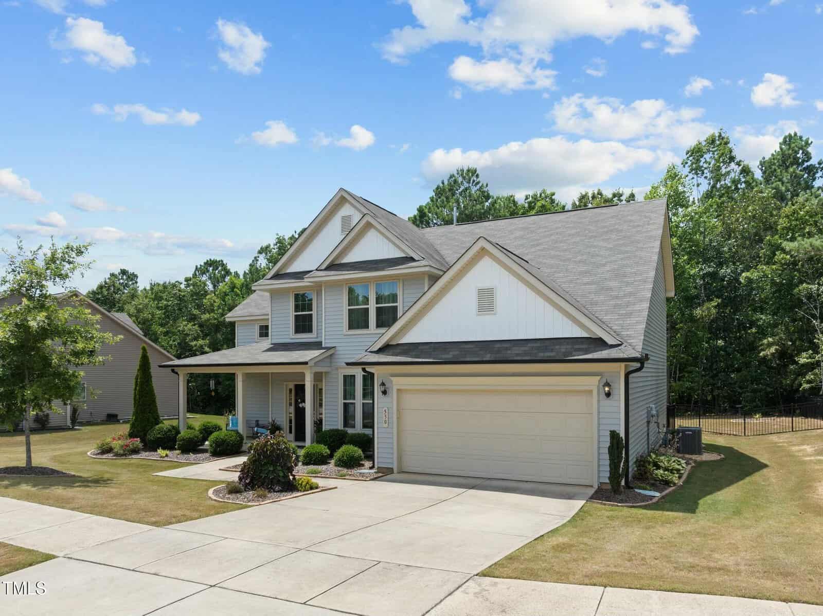 Two-story suburban house with light-colored siding, a covered front porch, two-car garage, and neatly landscaped front yard. The home sits on a corner lot with a concrete driveway and surrounded by trees under a blue sky.