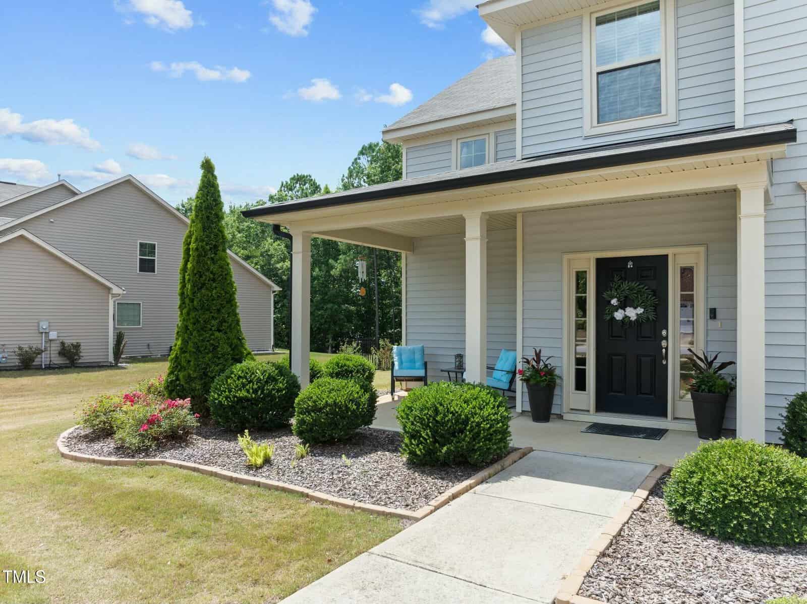 A modern house with light gray siding, a covered front porch, black door, two blue chairs, potted plants, and neatly trimmed bushes lining a walkway on a sunny day.