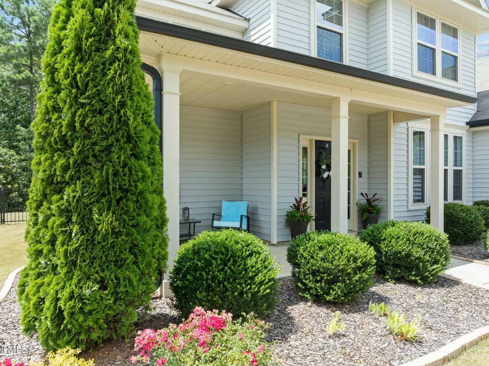 A tidy house with light gray siding, black shutters, and a covered front porch featuring a black door, potted plants, a turquoise bench, trimmed bushes, and vibrant flowers in the foreground.
