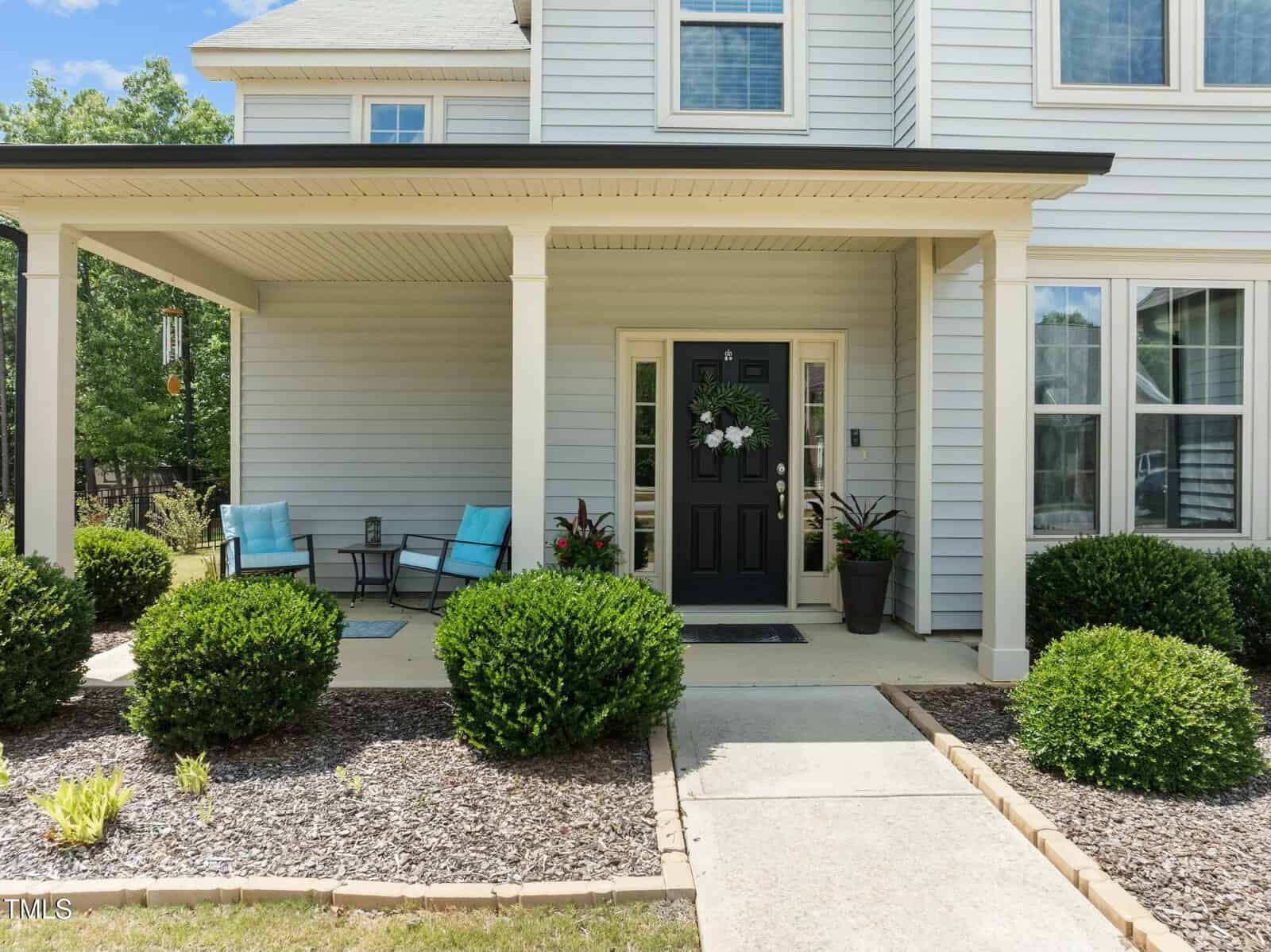 Front view of a modern house with light siding, a covered porch, black front door, two blue chairs, small table, green bushes, and a concrete walkway leading to the entrance.