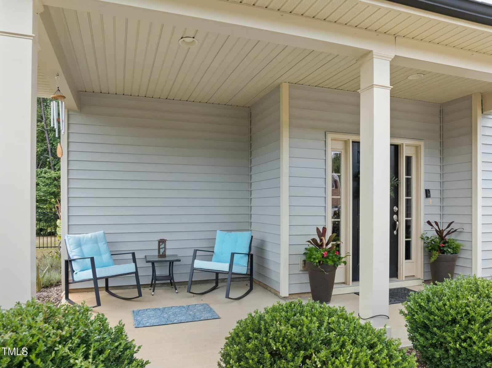 Covered porch with two light blue cushioned chairs, small table, potted plants, a doormat, and a wind chime, in front of a light gray house with double glass doors.