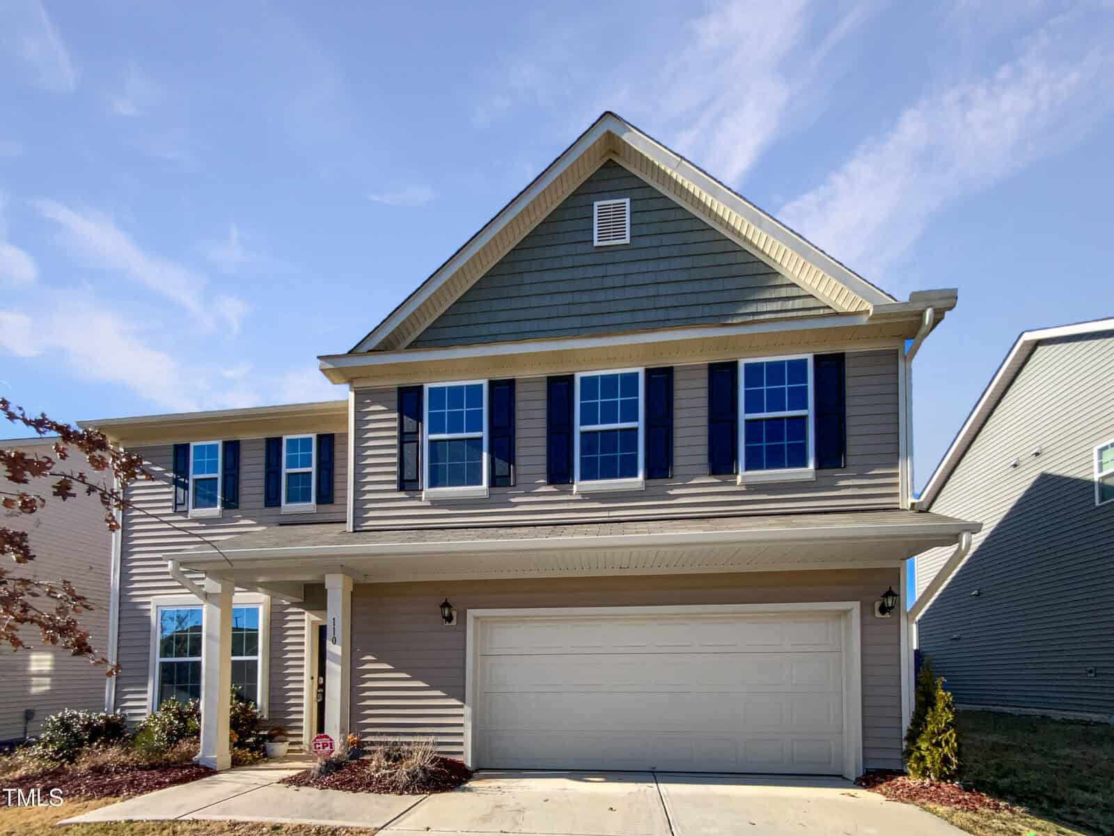 A two-story suburban house with tan siding, dark blue shutters, multiple windows, and a double garage. The home has a covered entryway and minimal landscaping, under a clear blue sky.