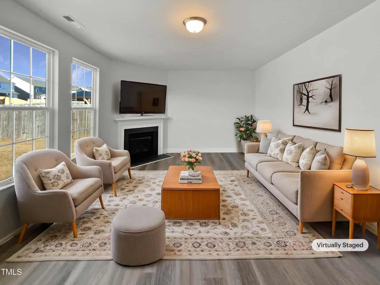 A living room with a large window, beige armchairs, a tan sofa, a wooden coffee table, an area rug, and a flat-screen TV mounted above a fireplace. A painting, lamp, and plant complete the virtually staged space.