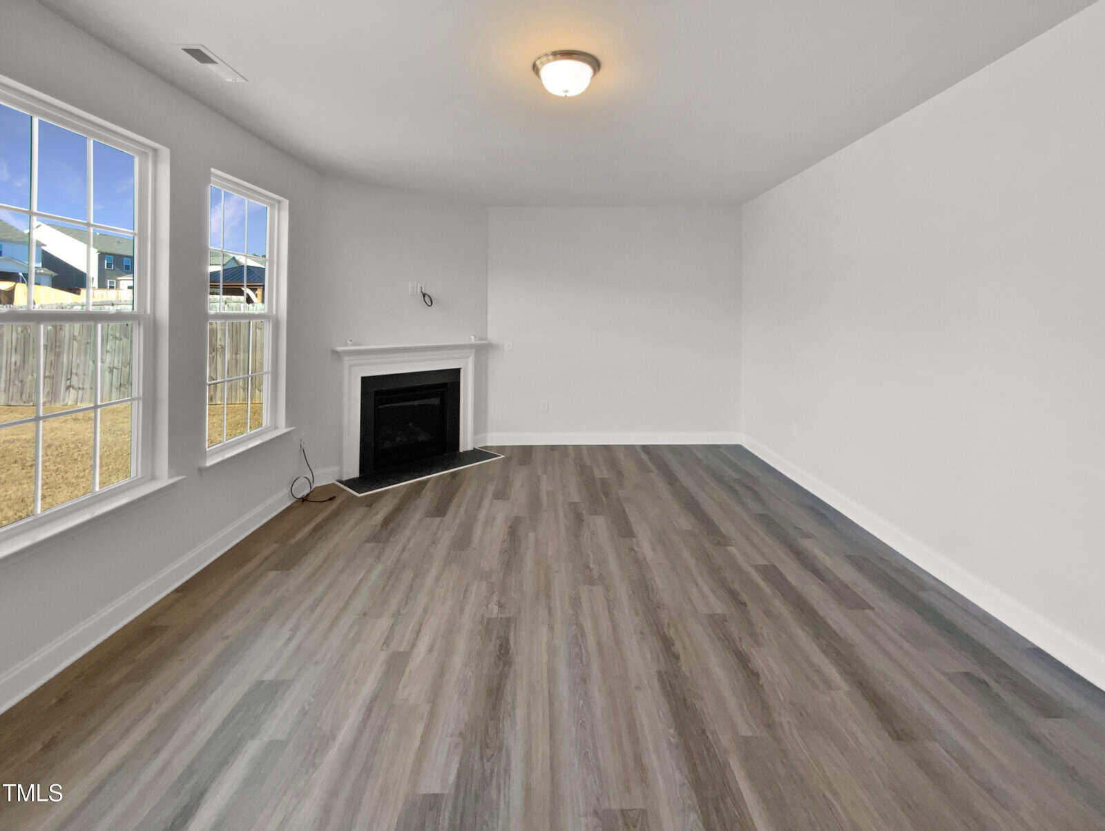 Unfurnished living room with gray wood-look flooring, white walls, a ceiling light fixture, a fireplace in the corner, and two large windows overlooking a fenced yard.