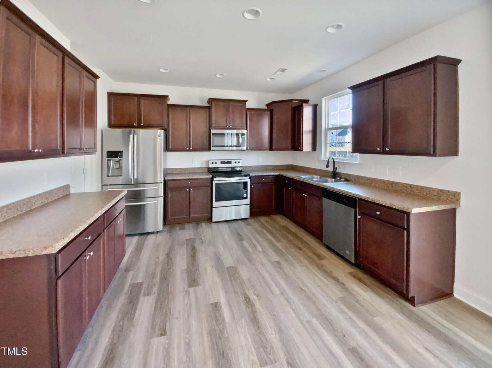 Modern kitchen with dark wood cabinets, stainless steel appliances, light brown countertops, and wood-look flooring. A window above the sink lets in natural light. The kitchen is empty and clean.