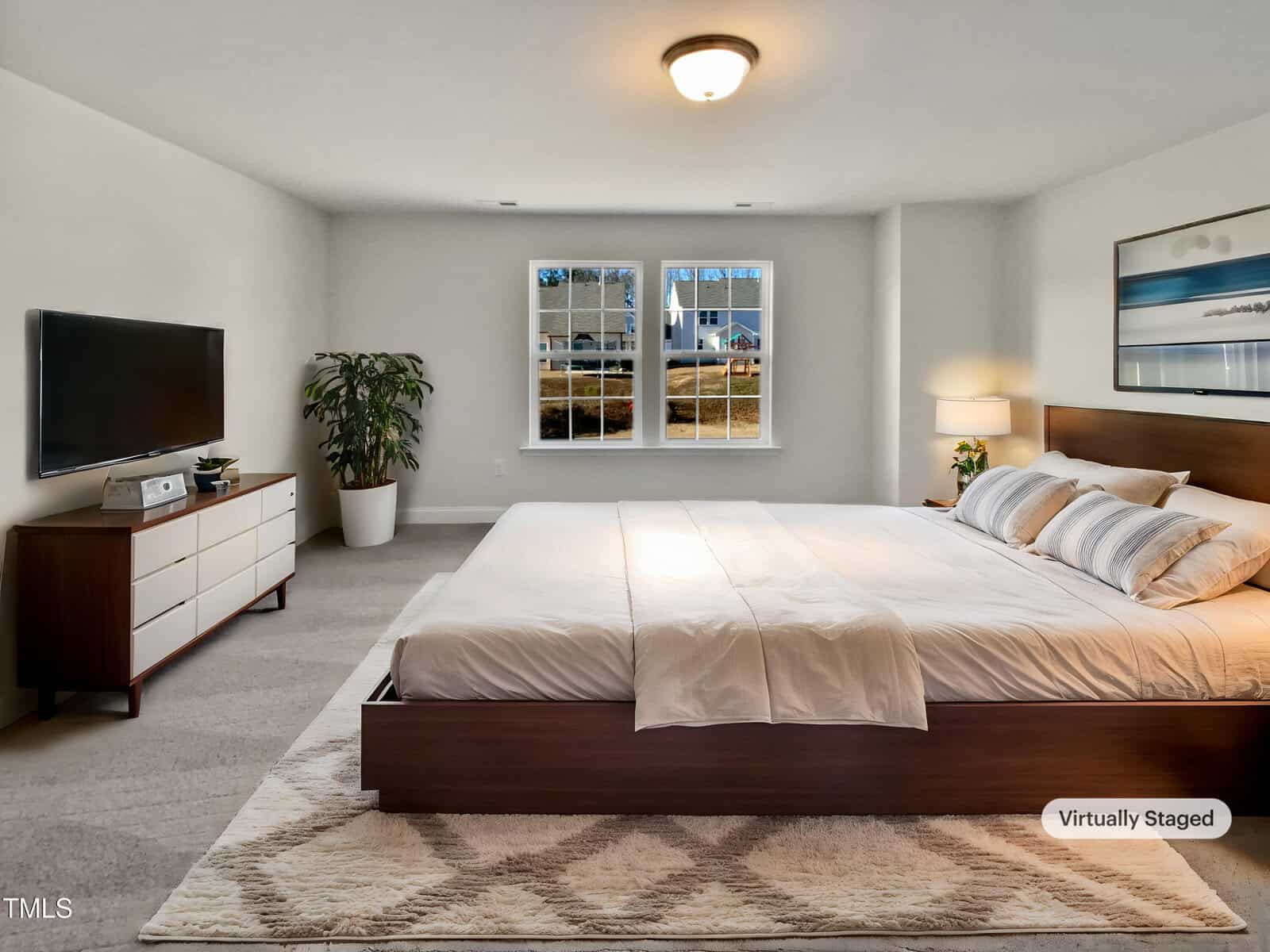 A spacious bedroom with a large bed, two windows, a potted plant, a dresser with a TV, a nightstand with a lamp, and neutral-colored walls and carpet. The image is labeled as virtually staged.