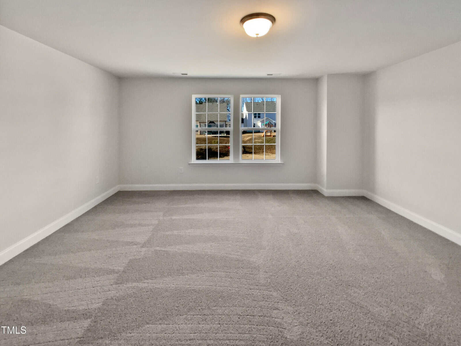 A spacious, empty room with light gray carpet, white walls, a ceiling light fixture, and two large windows showing a view of a street and houses outside.