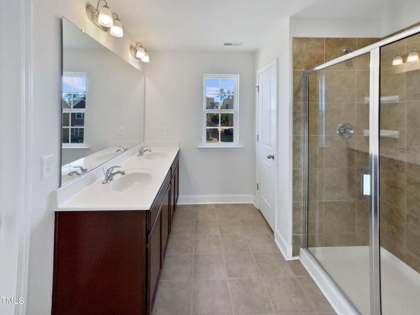 Modern bathroom with double sinks, a large mirror, tile floor, and a glass-enclosed shower with tan tiles. A window lets in natural light. The walls are white, and cabinetry under the sinks is dark wood.