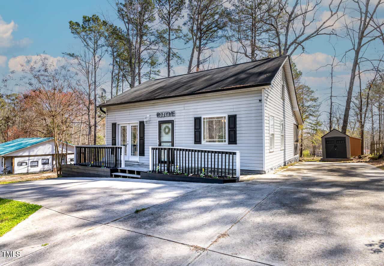 A small, single-story white house with black trim, front porch, and sloped roof sits on a concrete driveway surrounded by trees. A small shed and another building are visible in the background.