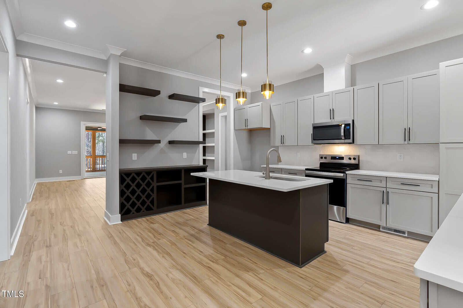 Modern kitchen with light wood floors, gray cabinets, a center island with sink, stainless steel appliances, built-in open shelving, wine rack, and three pendant lights hanging above the island.