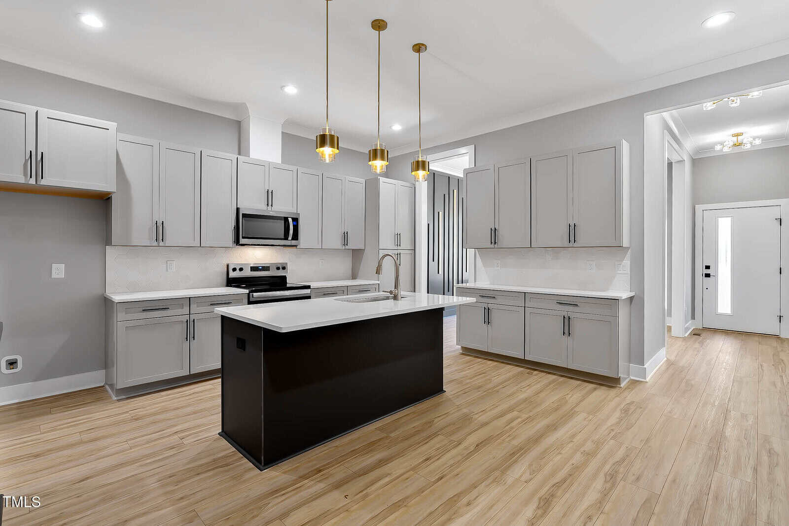 Modern kitchen with light gray cabinets, stainless steel appliances, a black island with a white countertop, three gold pendant lights, light wood flooring, and white walls. A hallway and door are visible in the background.