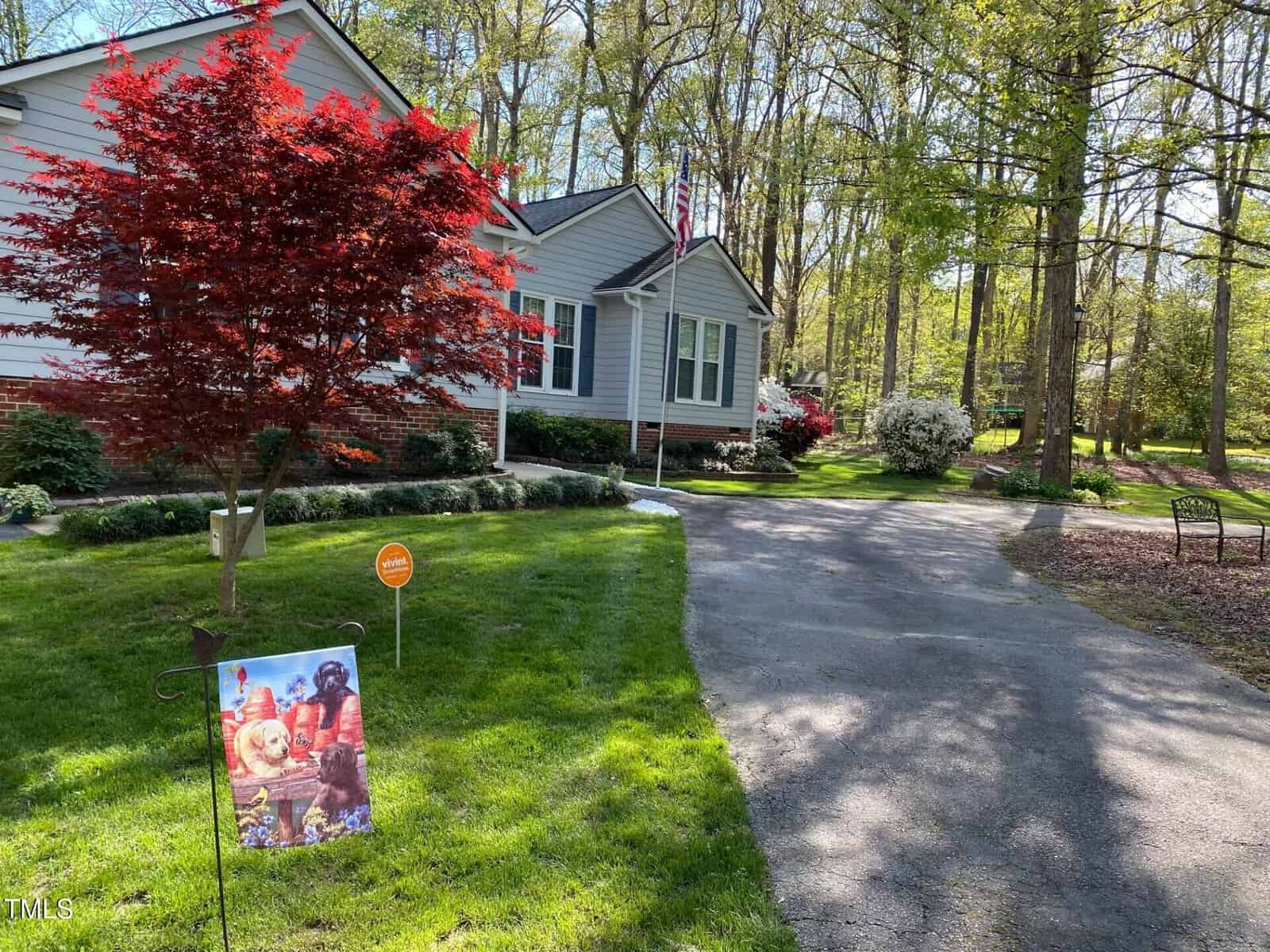 A light gray house with white trim sits beside a paved driveway lined with green grass, bushes, and trees. A small red-leafed tree and a decorative flag featuring dogs are visible in the front yard.