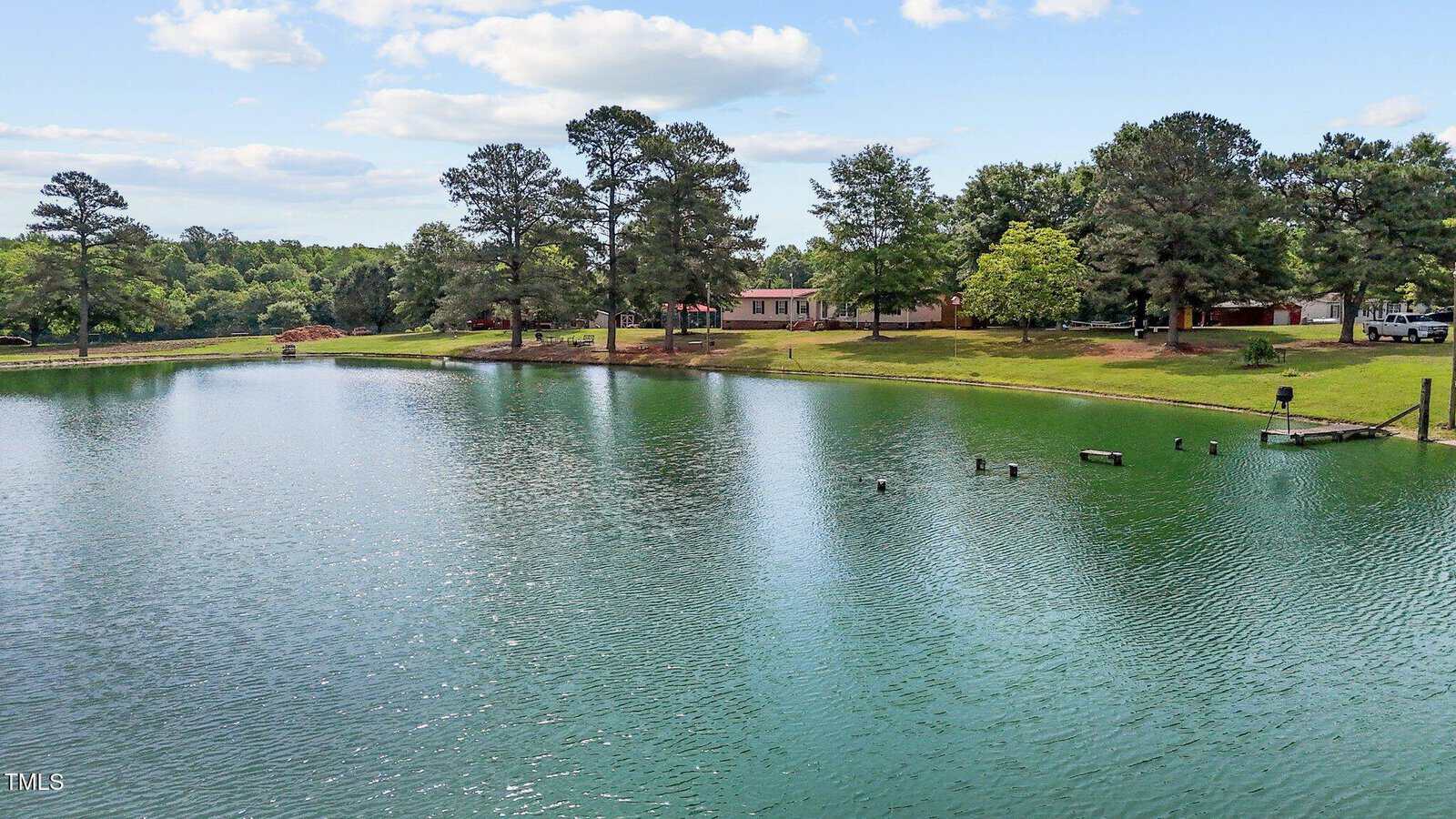 A pond with greenish water surrounded by trees and grassy banks; several buildings are visible among the trees under a partly cloudy sky. Wooden posts and a small dock extend into the water.