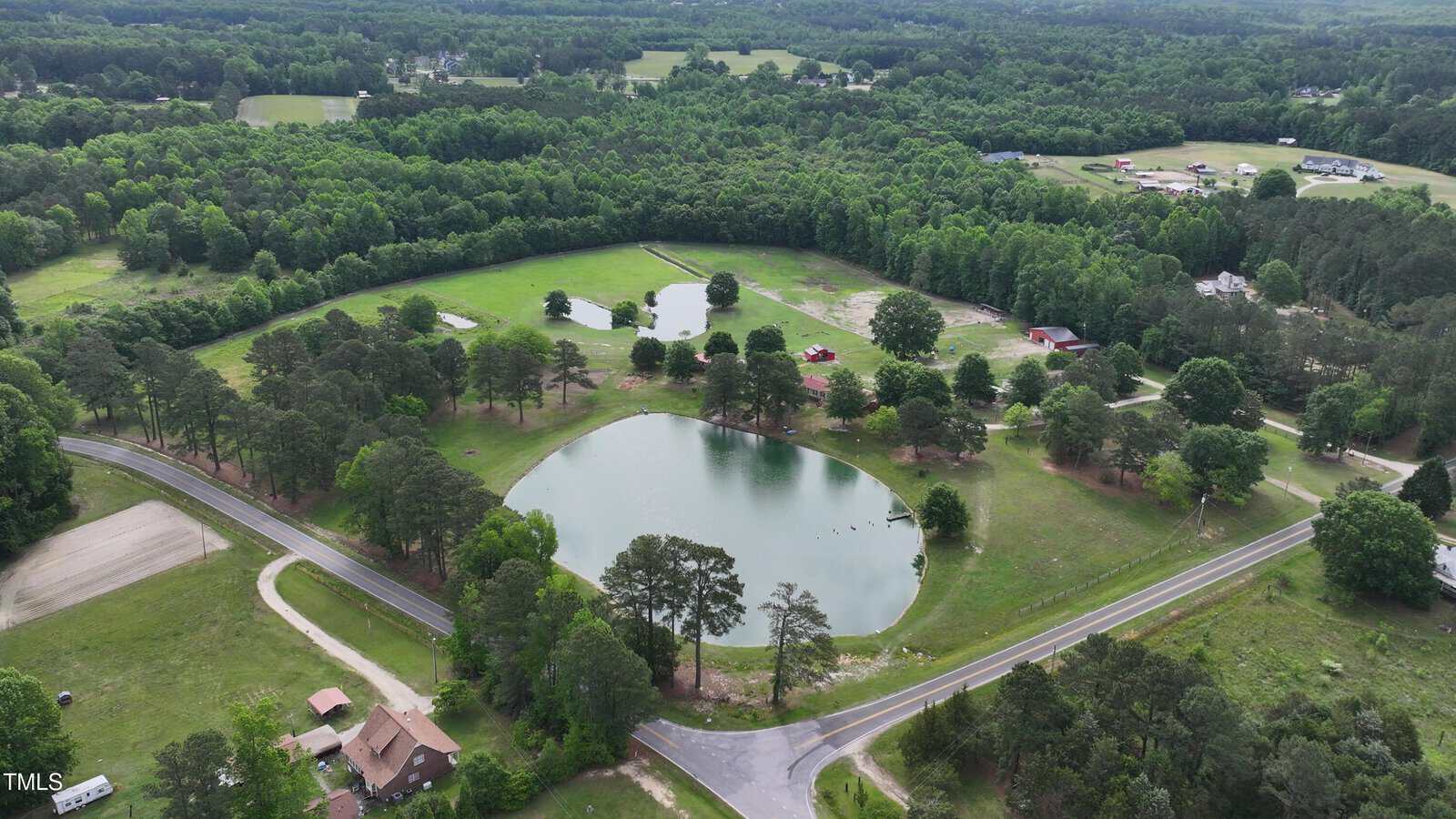 Aerial view of a rural area with two ponds surrounded by trees and grassy fields, intersected by paved and dirt roads, with scattered buildings and houses visible among the landscape.