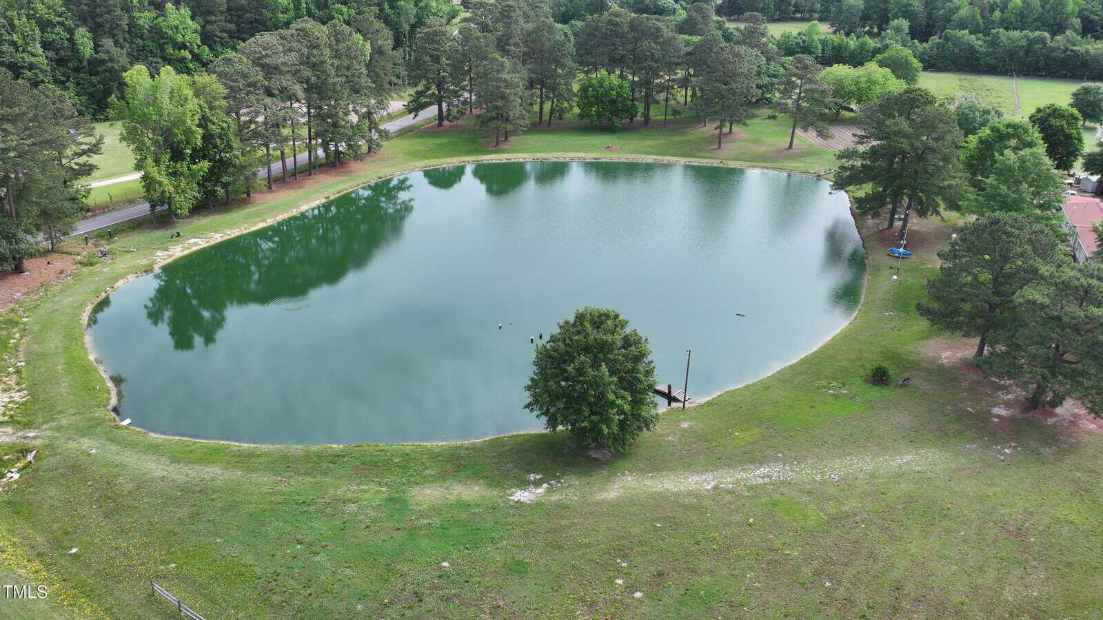 Aerial view of a small pond surrounded by grass and trees, with a dock extending into the water and a road visible in the background under a partly cloudy sky.