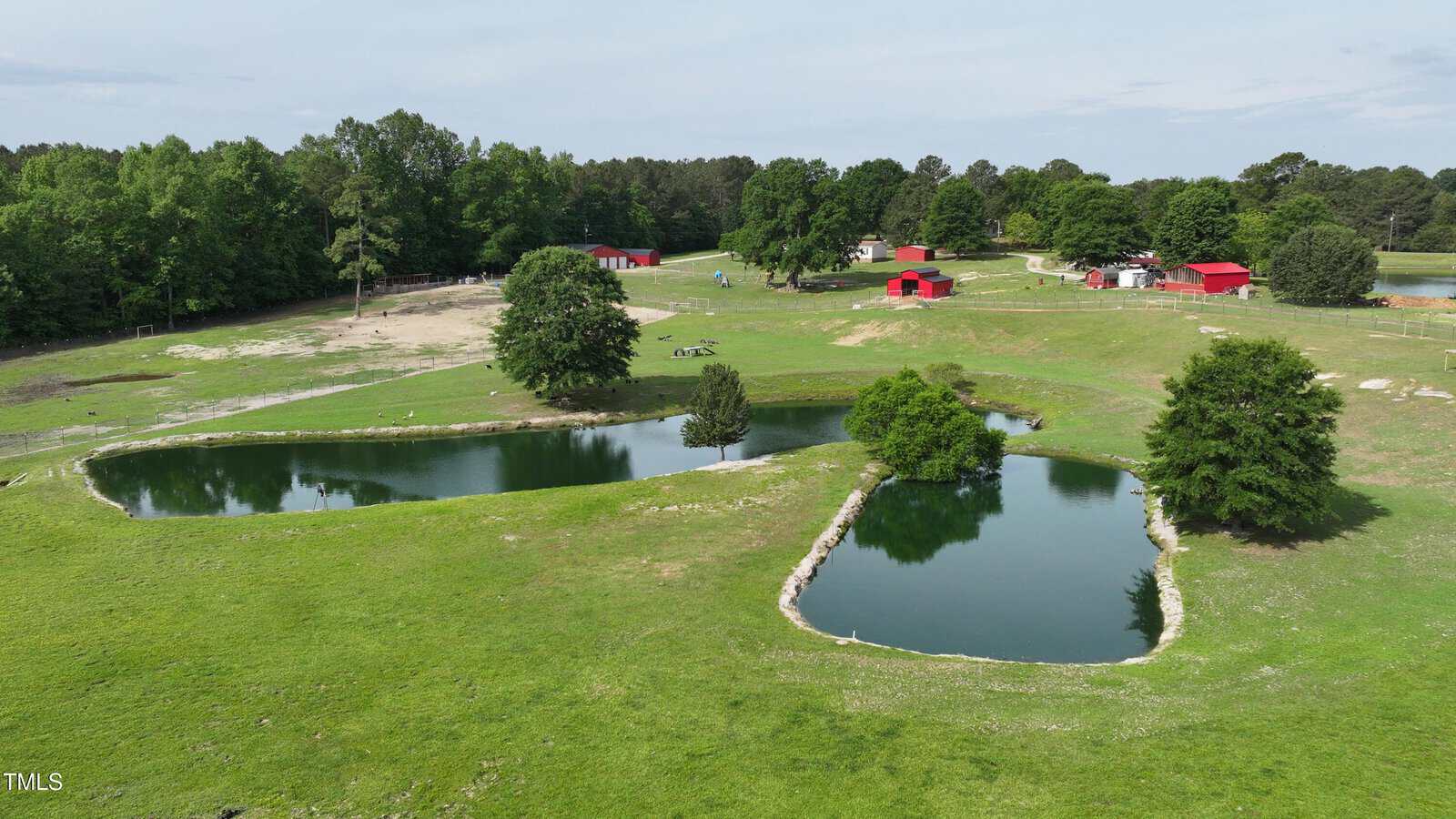 Aerial view of a grassy property with two small ponds, several large trees, and multiple red-roofed buildings in the background, surrounded by woods and open land under a partly cloudy sky.