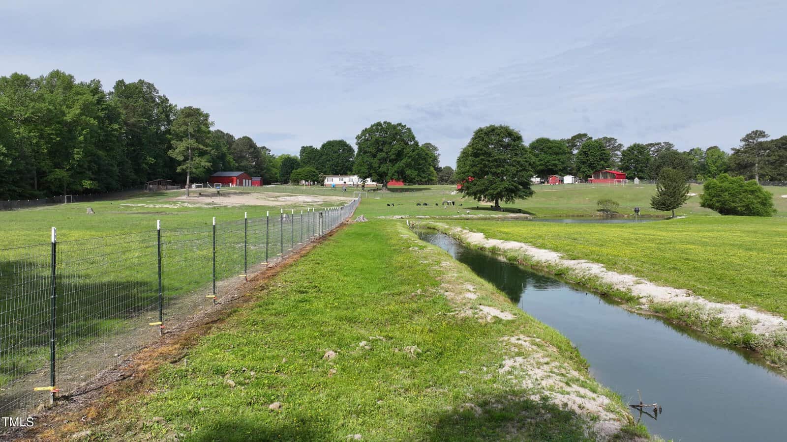 A wire fence runs beside a narrow stream through a grassy field, with trees and several red and white barns and buildings in the background under a partly cloudy sky.