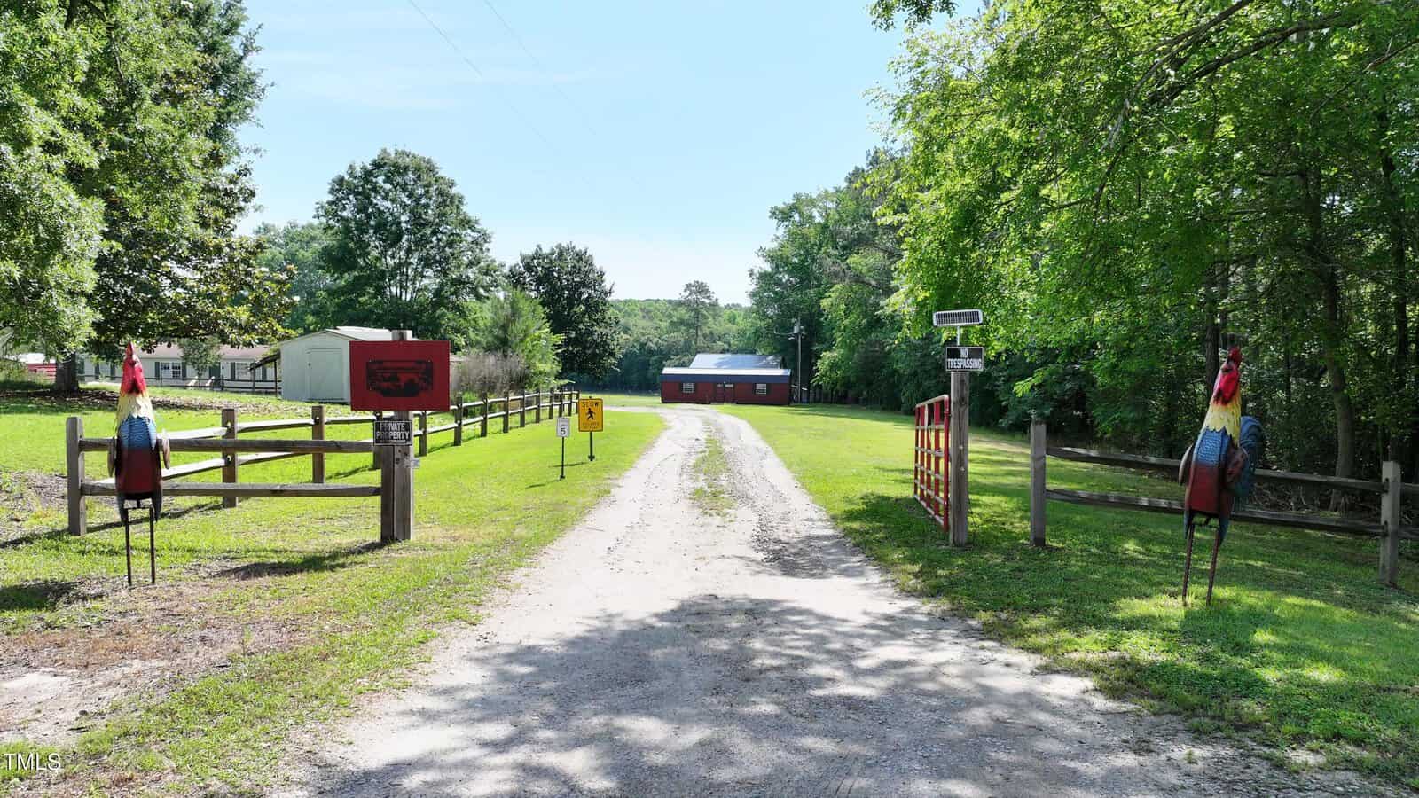 A gravel driveway leads to a red barn with a metal roof. There are fences, metal rooster statues on either side of the entrance, green grass, and trees surrounding the area. A sign and a gate are visible on the right.