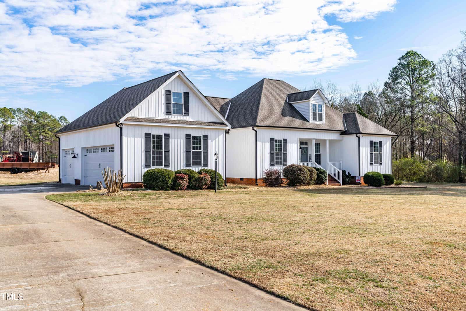 A white single-story house with gray roof shingles, a front porch with steps, two dormer windows, and an attached garage sits on a large lawn with a concrete driveway and is surrounded by trees.