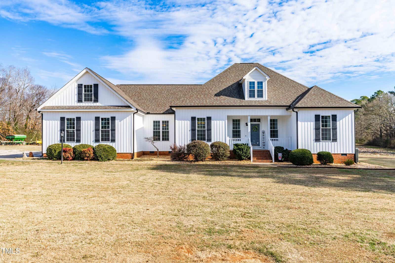 A white, single-story house with black shutters, a gabled roof, and a covered front porch. The house sits on a large lawn with some shrubs and trees in the background under a partly cloudy sky.