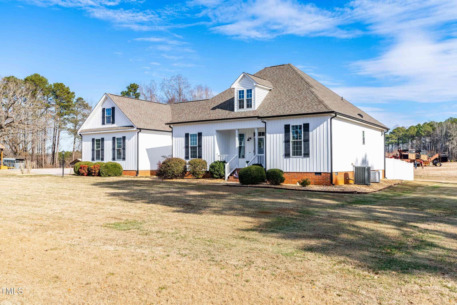 A white single-story house with dark shutters and a front porch sits on a large, grassy lot with minimal landscaping. There are trees and a clear blue sky in the background.