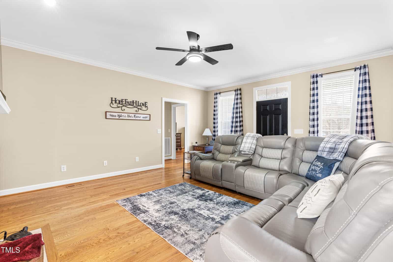 A cozy living room features a gray sectional sofa, rug, plaid curtains, wood floors, a ceiling fan, and a black front door. Wall art and a side table with a lamp add decorative touches.