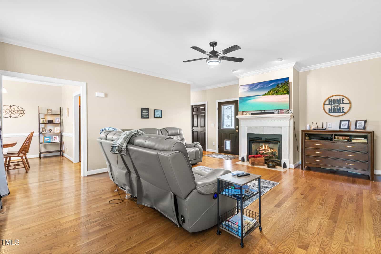 A living room with hardwood floors, two gray recliners, a ceiling fan, a fireplace with a TV above it, a dresser, and decorative items. An open doorway leads to a dining area with a wooden table and chairs.