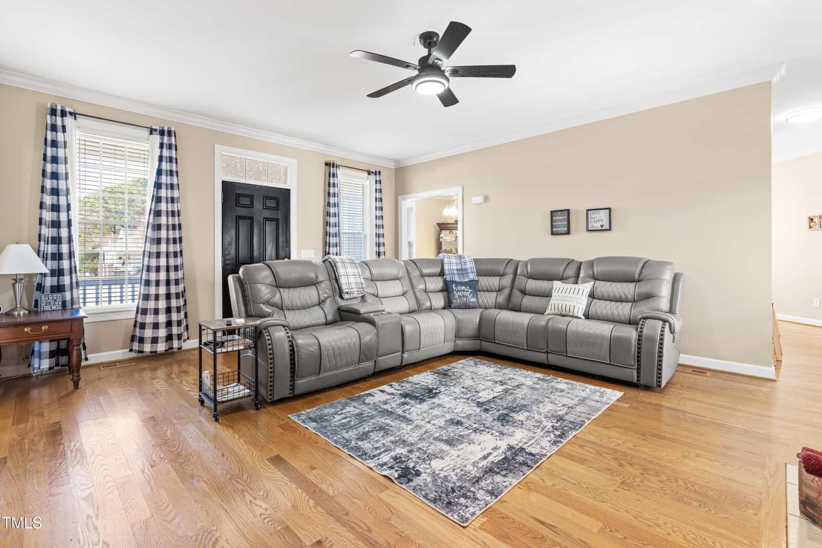A living room with a gray sectional sofa, patterned pillows, a rug, wooden floors, light tan walls, black front door, and two windows with blue plaid curtains. A ceiling fan and wall art complete the space.