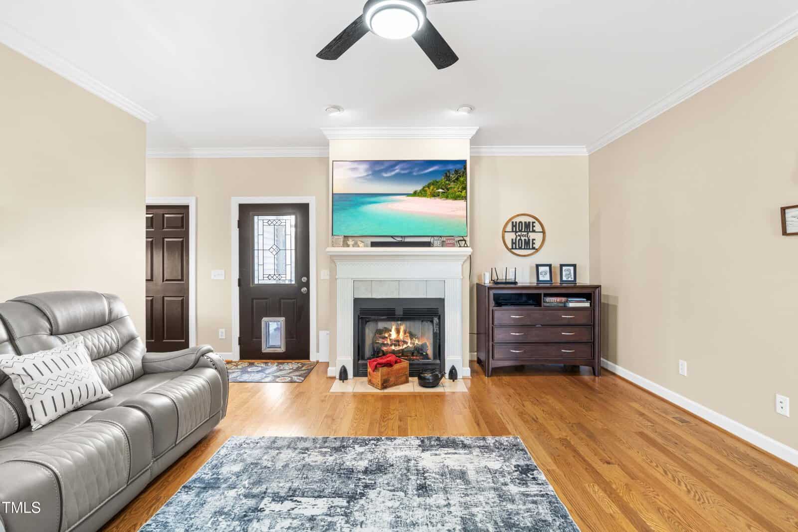 A bright living room with a gray sofa, a fireplace with a fire burning, a wall-mounted TV displaying a beach scene, wooden floors, a ceiling fan, and neutral walls.