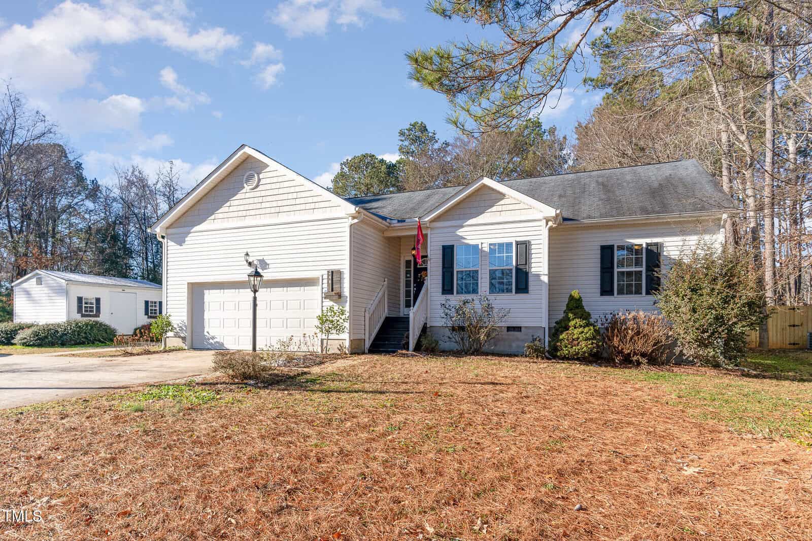 A single-story white house with an attached two-car garage and front steps, set on a dried grass lawn with some bushes. There is a smaller white outbuilding in the background and trees surrounding the property.