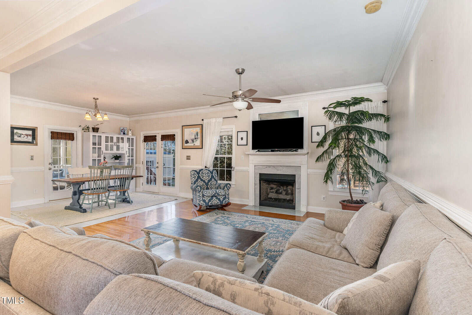 A spacious living room with a large beige sectional sofa, a coffee table, a fireplace with a TV mounted above it, a patterned armchair, a dining area in the background, and a potted plant by the fireplace.