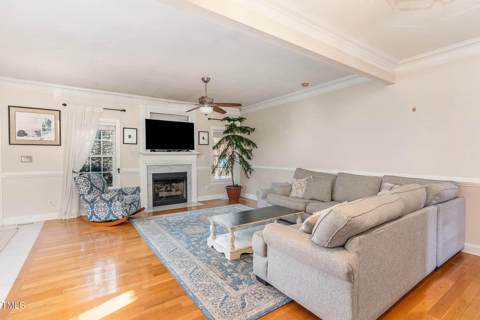 A living room with hardwood floors, a gray sectional sofa, a patterned armchair, a blue area rug, a coffee table, a fireplace with a TV above it, framed art, a floor plant, and a ceiling fan.