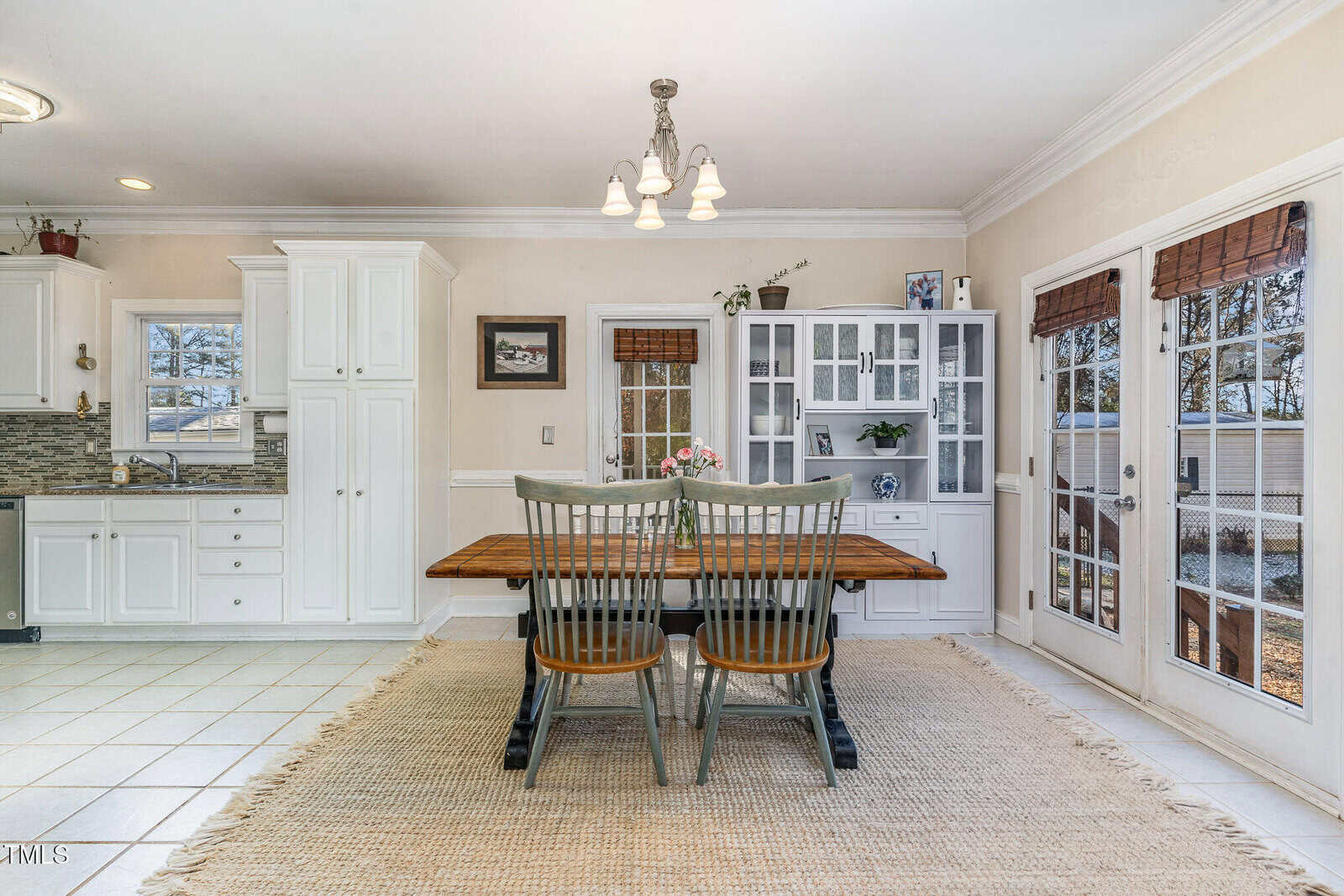 A bright kitchen and dining area with white cabinets, a wooden dining table with two chairs, a light fixture above, a woven rug, and glass doors leading outside. A vase with flowers is on the table.