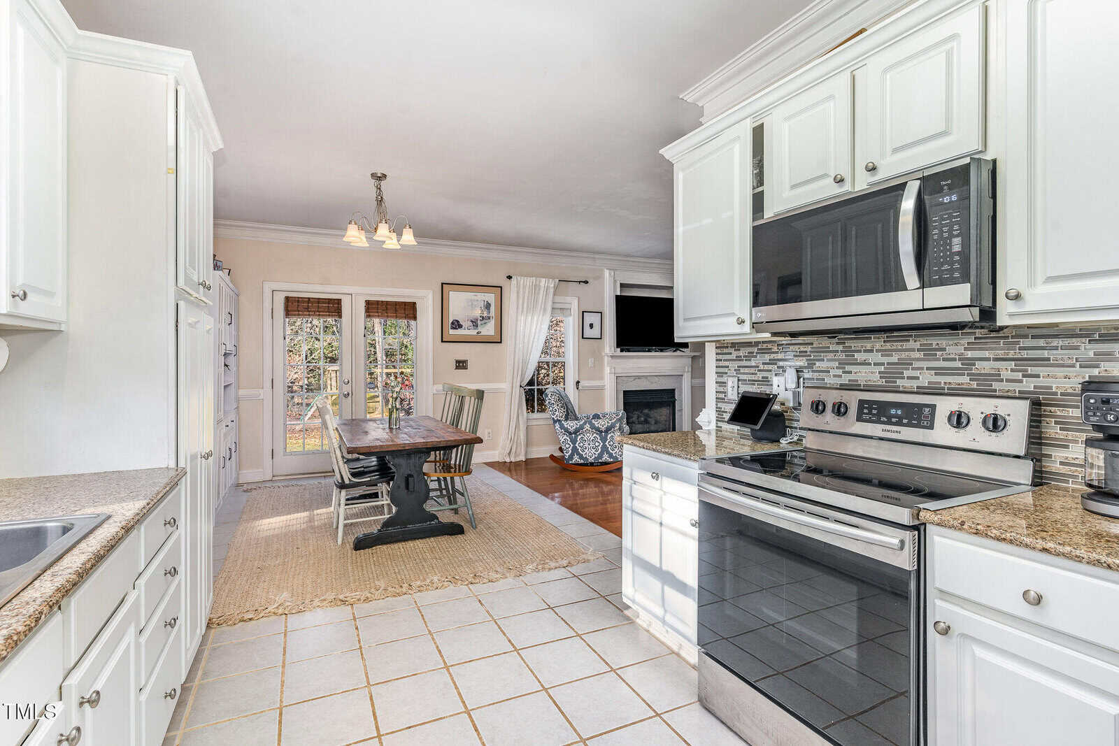 Bright kitchen with white cabinets, stainless steel appliances, and tile floor. A dining area with a wooden table and chairs sits near double glass doors. A living area with a fireplace and TV is visible in the background.