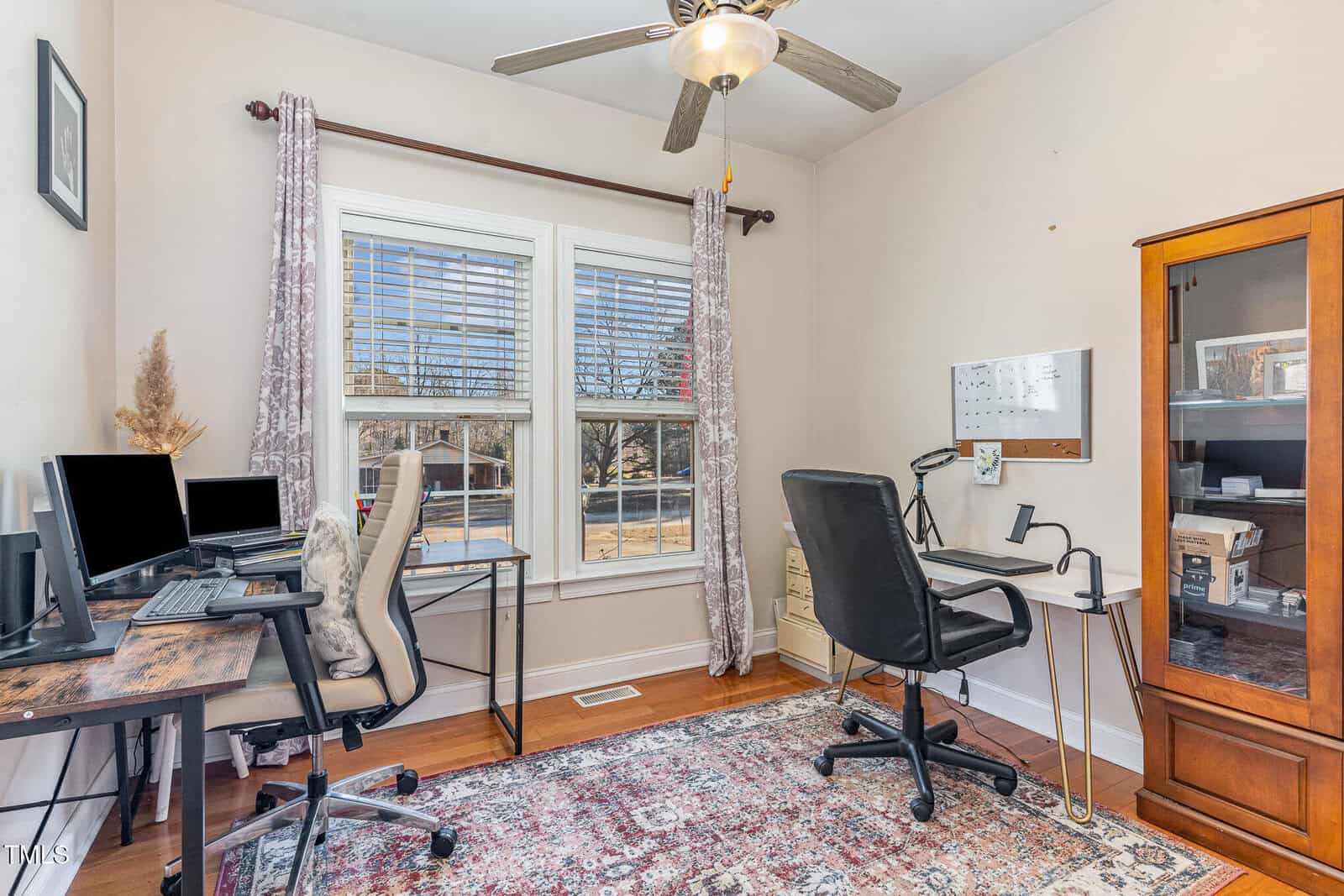 Bright home office with two desks, computers, a black chair, a glass cabinet, wall-mounted whiteboard, and large windows with patterned curtains letting in sunlight. Ceiling fan above, decorative rug on wooden floor.