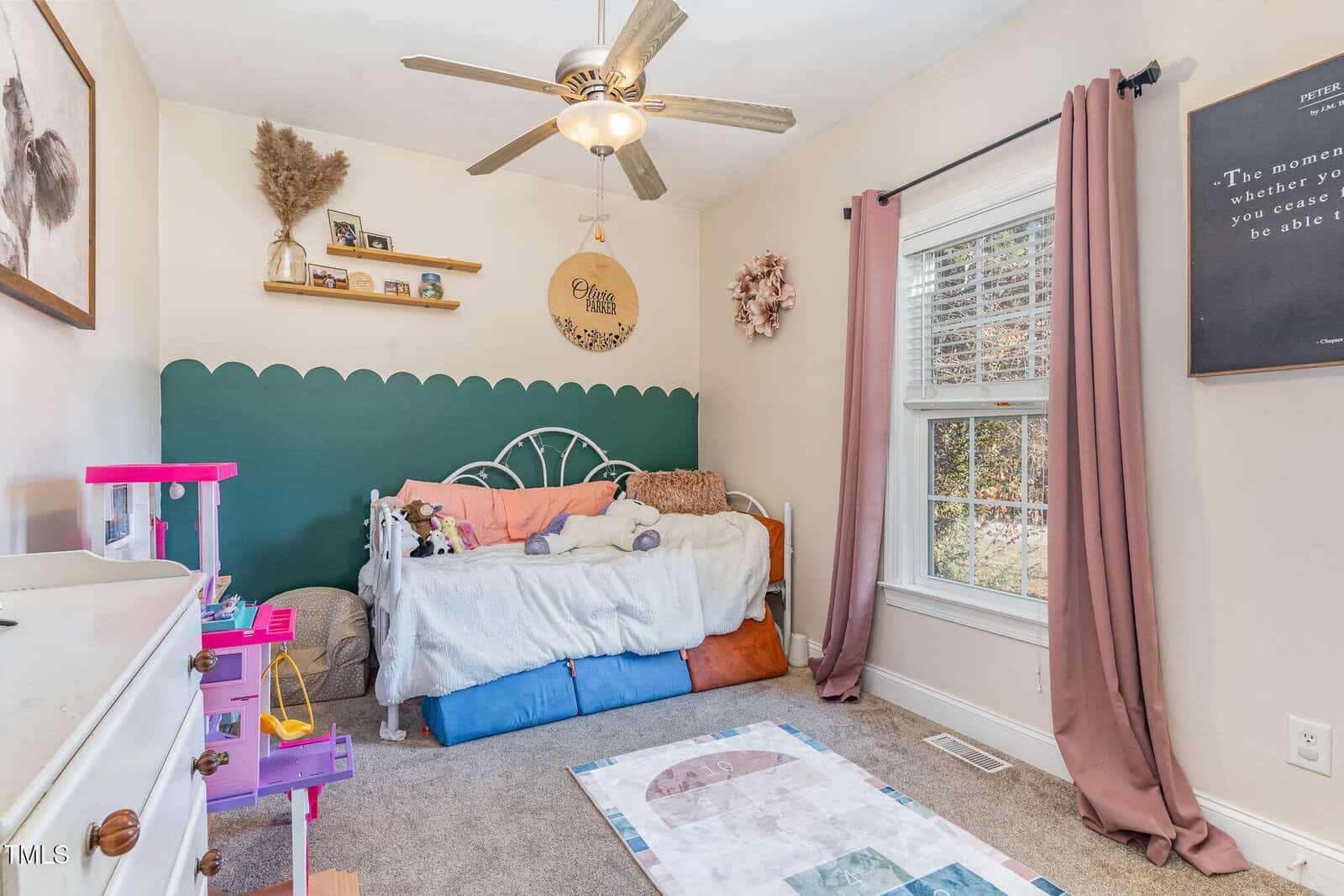 A cozy children’s bedroom with a white daybed, plush toys, a green scalloped accent wall, pink curtains, a ceiling fan, a rug, a dollhouse, and decorative shelves with art and pampas grass. Sunlight streams in through the window.