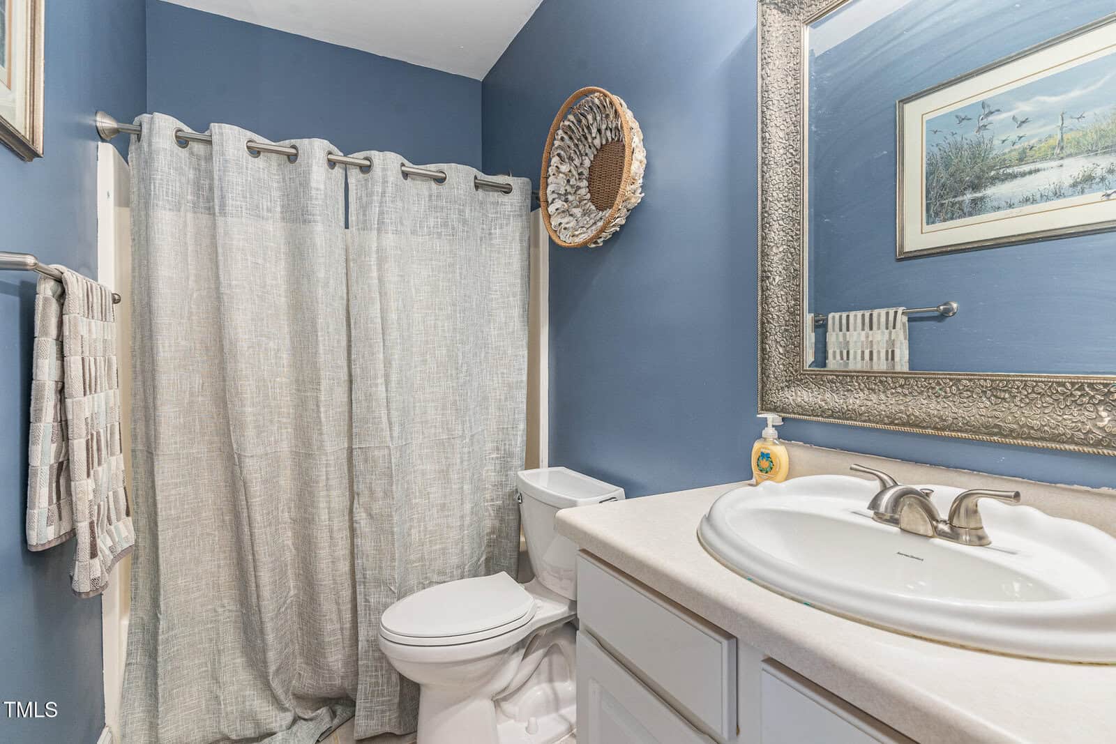 A bathroom with blue walls, a white sink and countertop, a toilet, a shower with a gray curtain, a towel rack with towels, a round decorative wall piece, and a framed landscape picture above the towel rack.