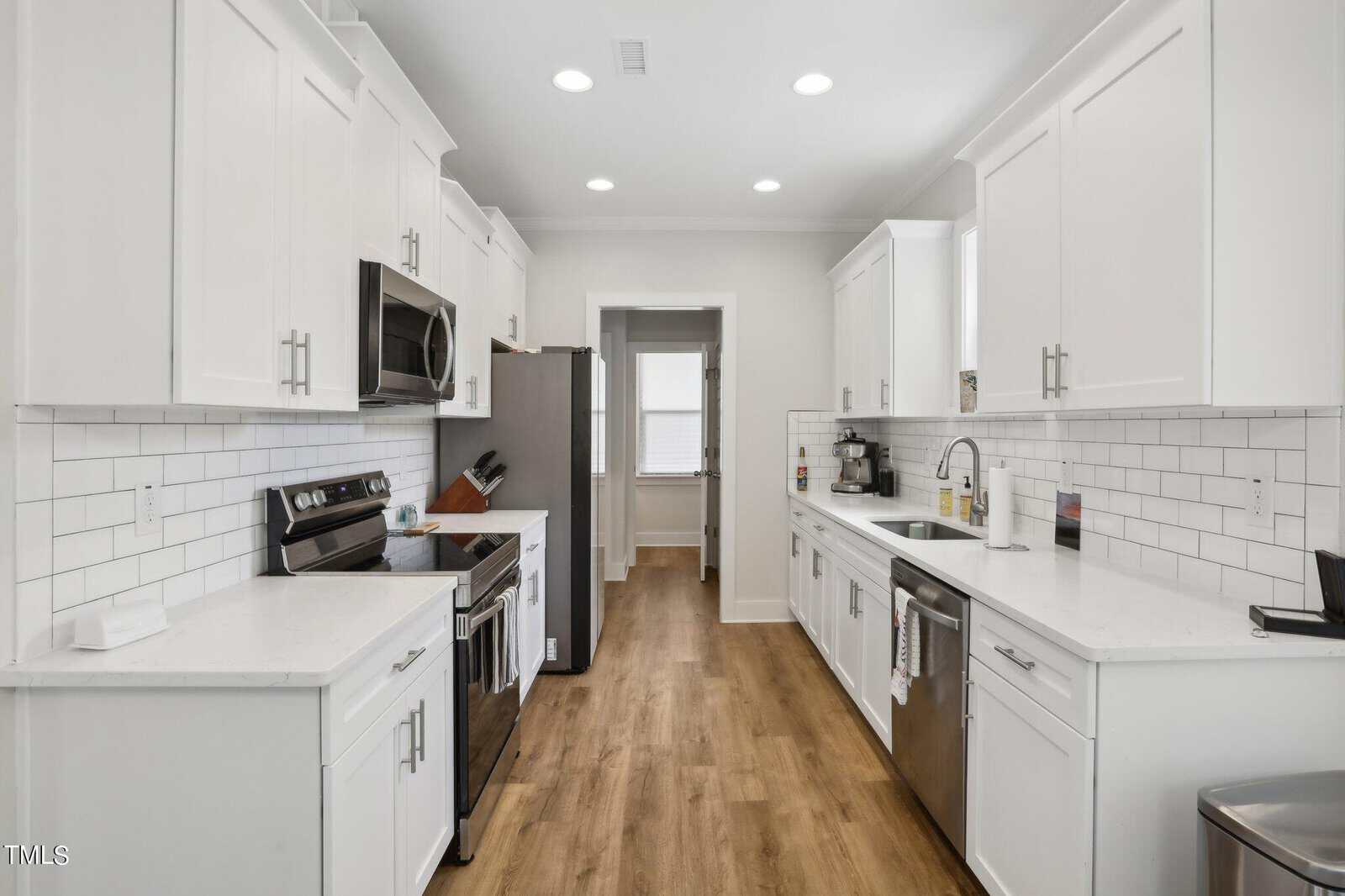A modern galley kitchen features white cabinets, white subway tile backsplash, stainless steel appliances, wood flooring, and recessed ceiling lights. A window is visible at the far end of the room.