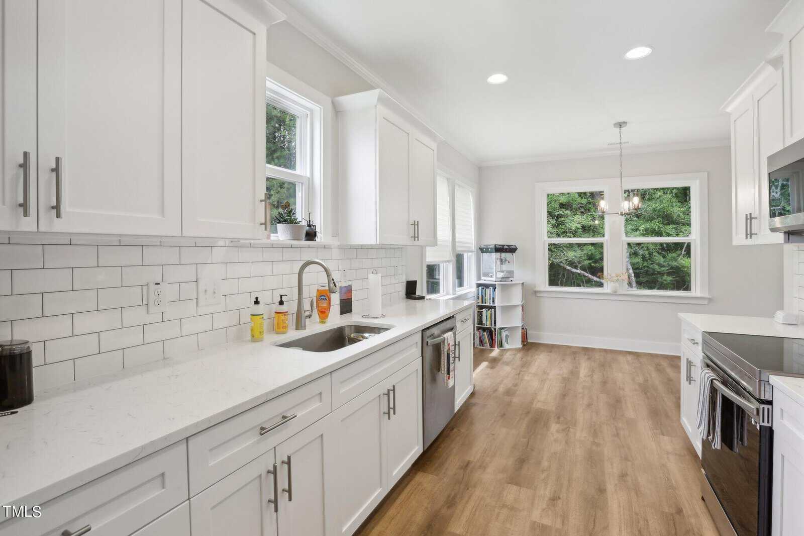 A modern kitchen with white cabinets, a white subway tile backsplash, stainless steel appliances, and wood-look flooring. Two windows above the sink and three more near a dining nook provide natural light.