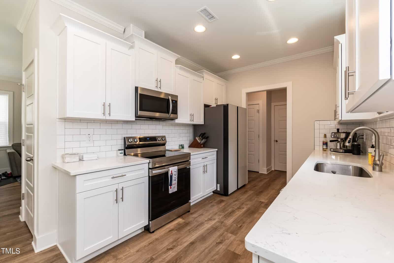 Modern kitchen with white cabinets, stainless steel appliances, subway tile backsplash, wood flooring, and quartz countertops. A microwave is mounted above the stove, and a refrigerator is next to the pantry door.