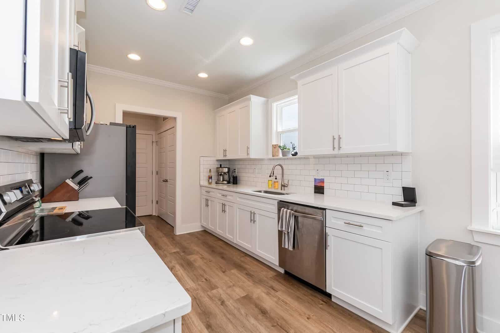 A modern kitchen with white cabinets, stainless steel appliances, white subway tile backsplash, wooden floor, and a window above the sink. A towel hangs on the dishwasher handle, and small appliances sit on the counter.